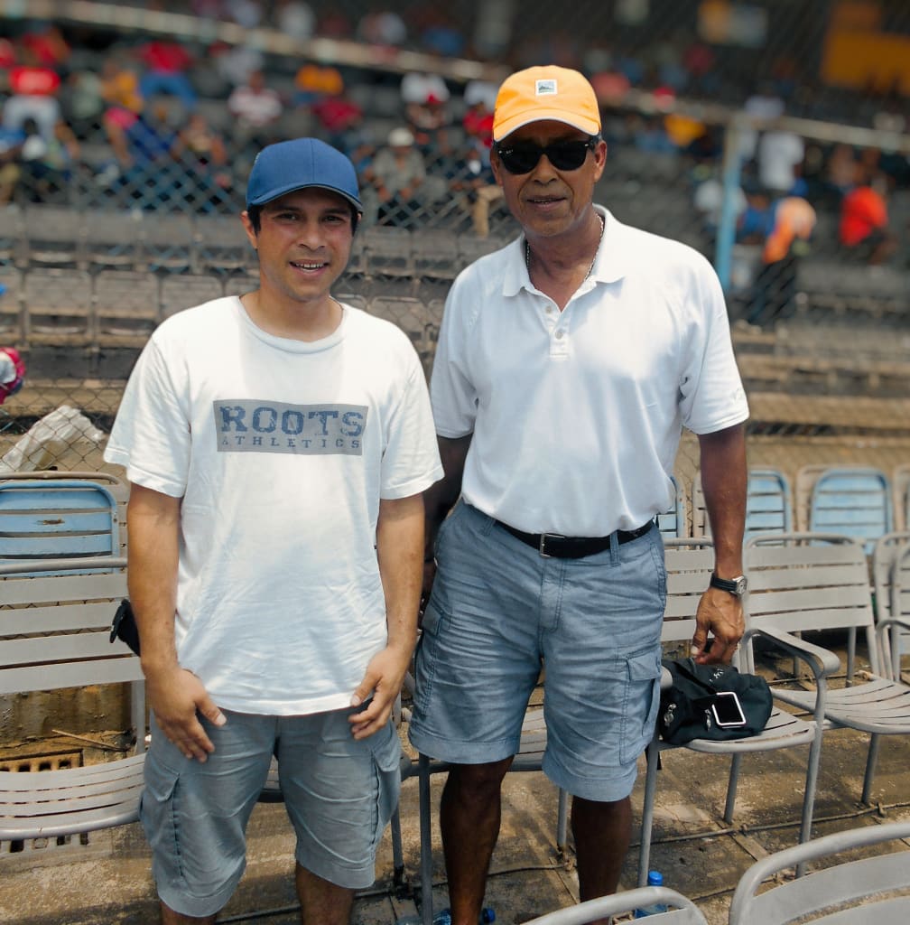 Two men in baseball caps and white t-shirts smile at the camera