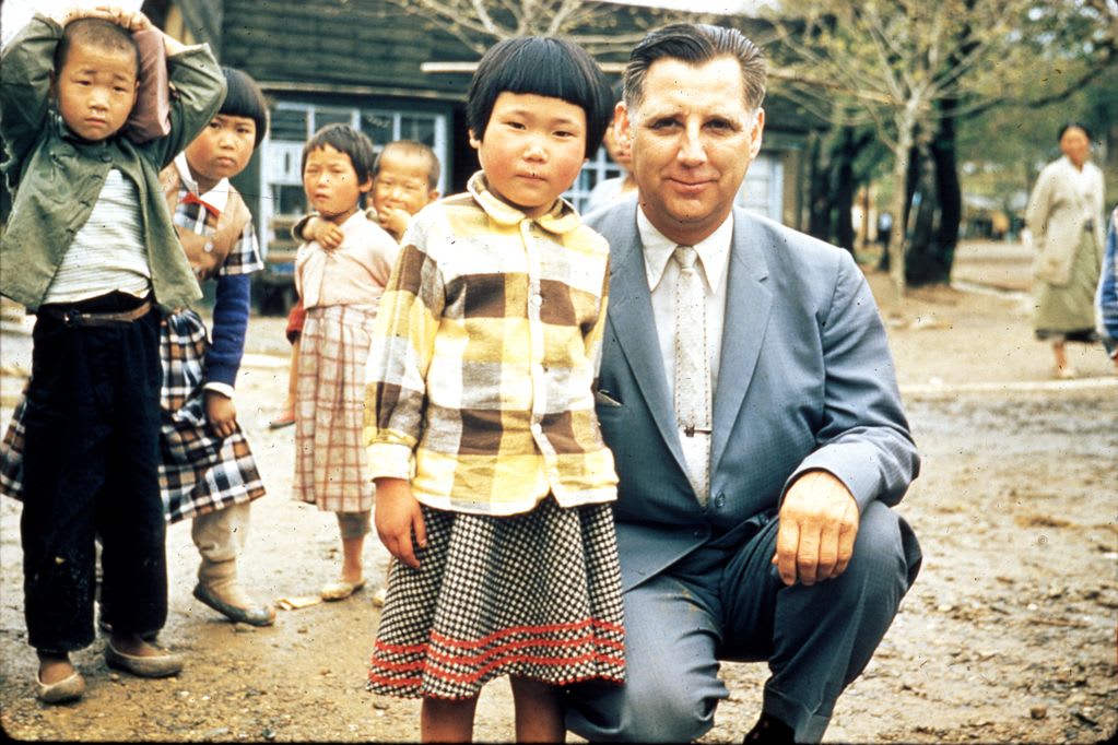 A man in a suit stands beside a young girl wearing a dress and a button up flannel shirt. There are kids in the background playing.
