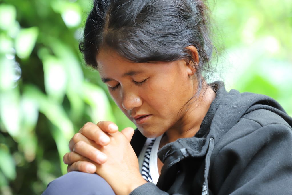 A woman in a black coat clasps her hands while closing her eyes and praying.