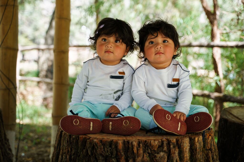 Twin boys dressed in blue pants and white shirts with thick dark hair sit together on a tree stump/