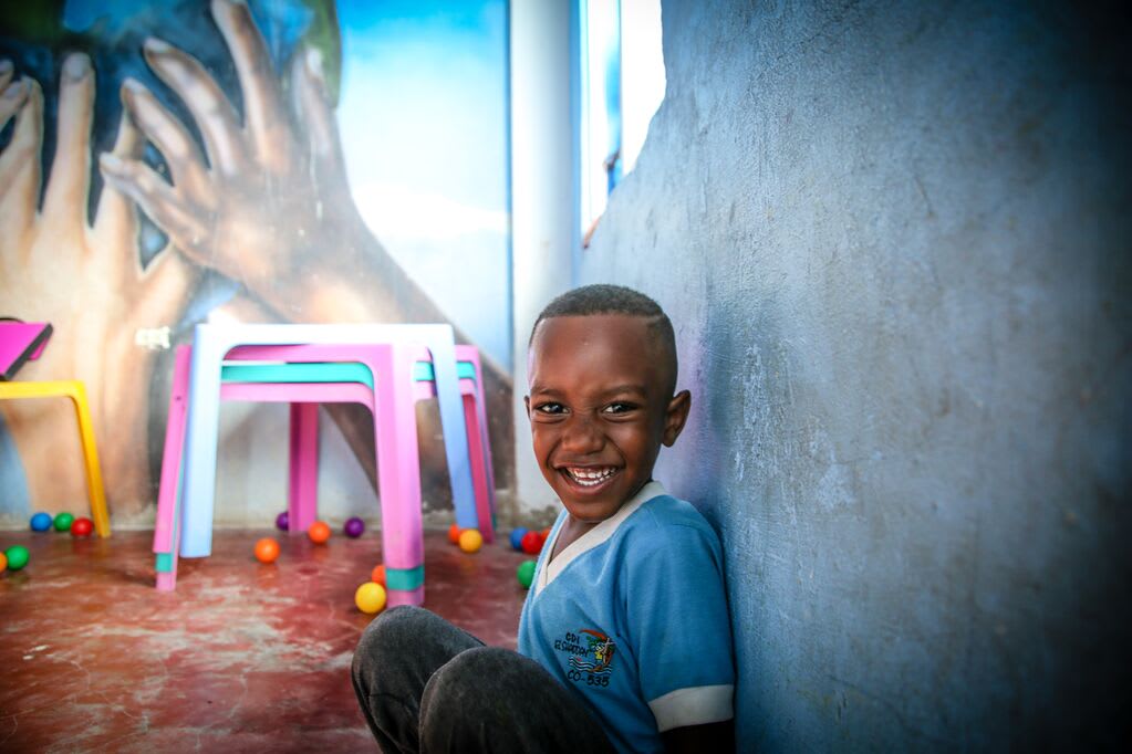 Little boy in a blue shirt sits against the wall of his Compassion centre and beams.