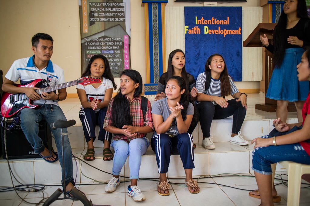 A group of 5 girls sits on the steps of their Compassion centre singing along to someone playing guitar
