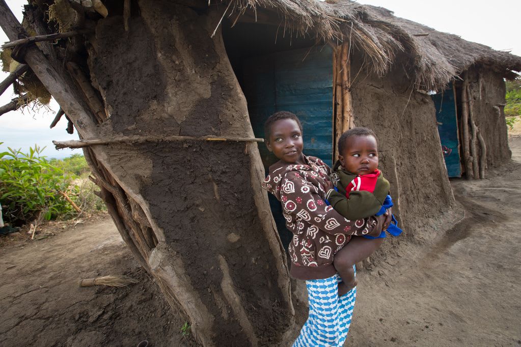 A young girl holds a baby in her arms while leaning against a home made from mud and sticks with a thatched roof.