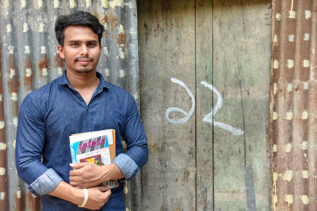 An adolescent male, teenager, Sujon wearing a blue denim shirt is holding several of his books. He is standing in front of a corrugated metal wall, building and a wooden doorway.