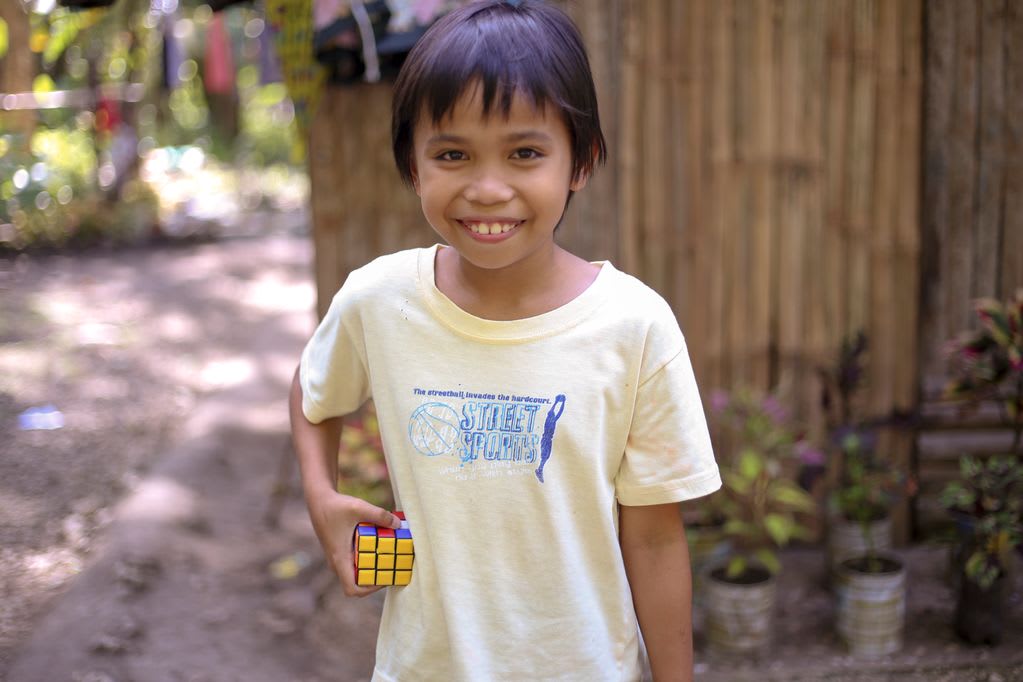 A smiling 10 year-old boy, male child, Jessie Mhar Villaplana, wearing a yellow shirt, is holding his Rubik’s cube. He is standing in front of a wooden, bamboo wall, fence and several potted plants.