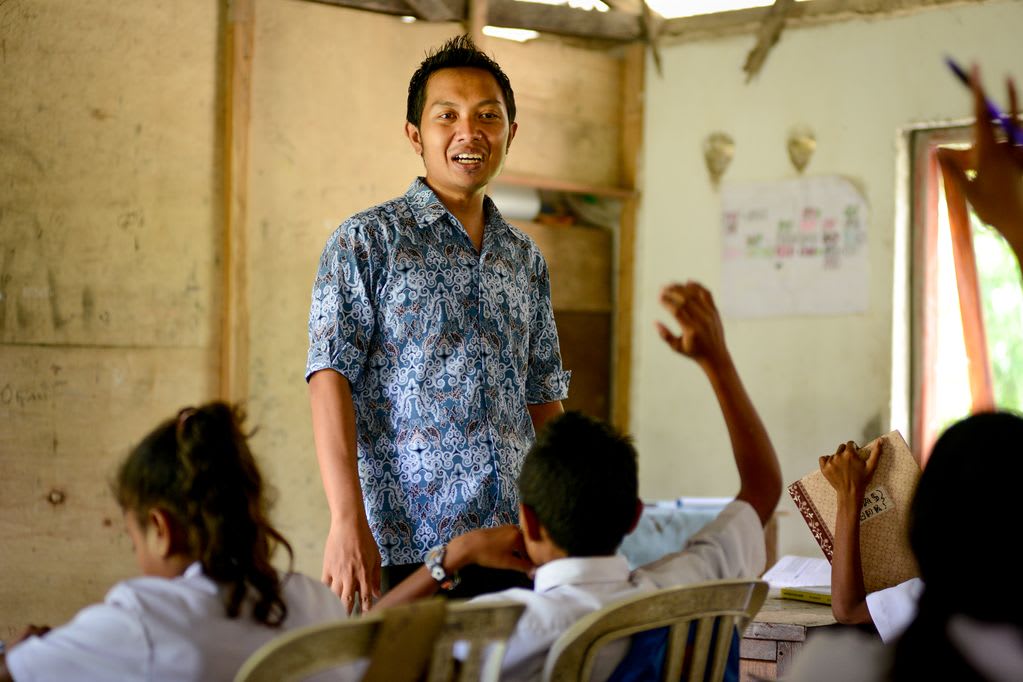 A man stands in a classroom made from plywood wearing a blue shirt, in front of children sitting in chairs raising their hands