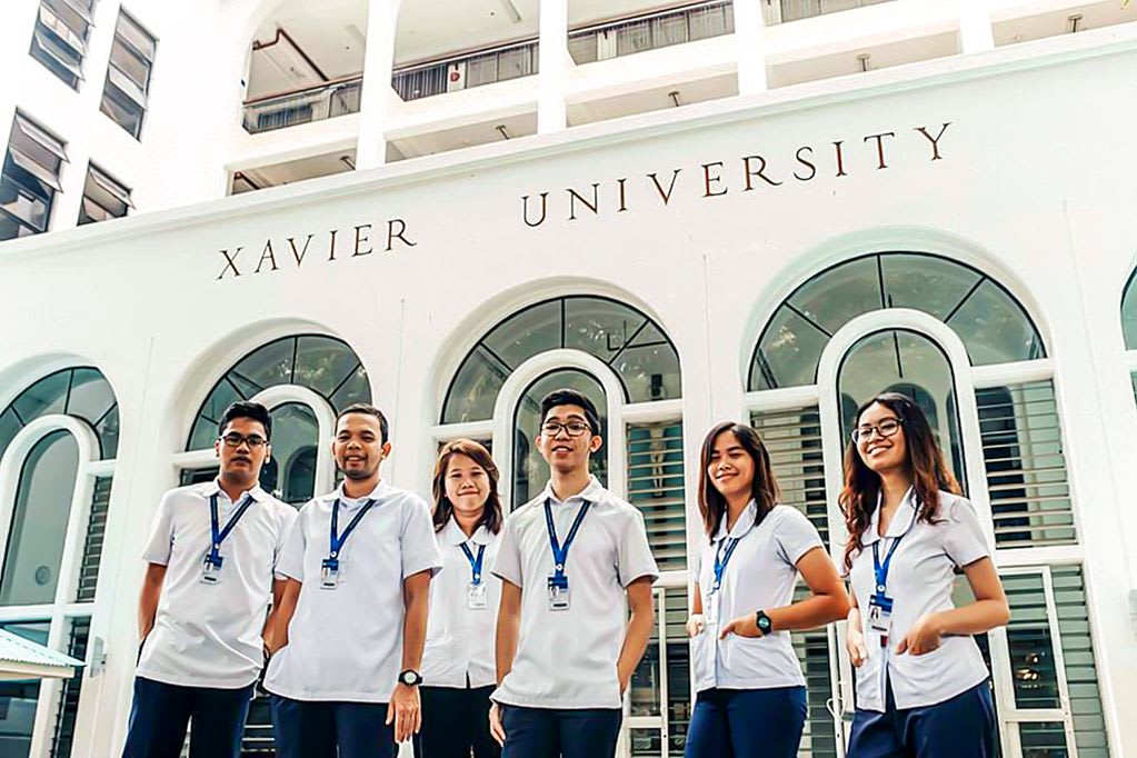 A group of six students stand outside wearing matching white shirts in front of a white building that is labeled "Xavier University"