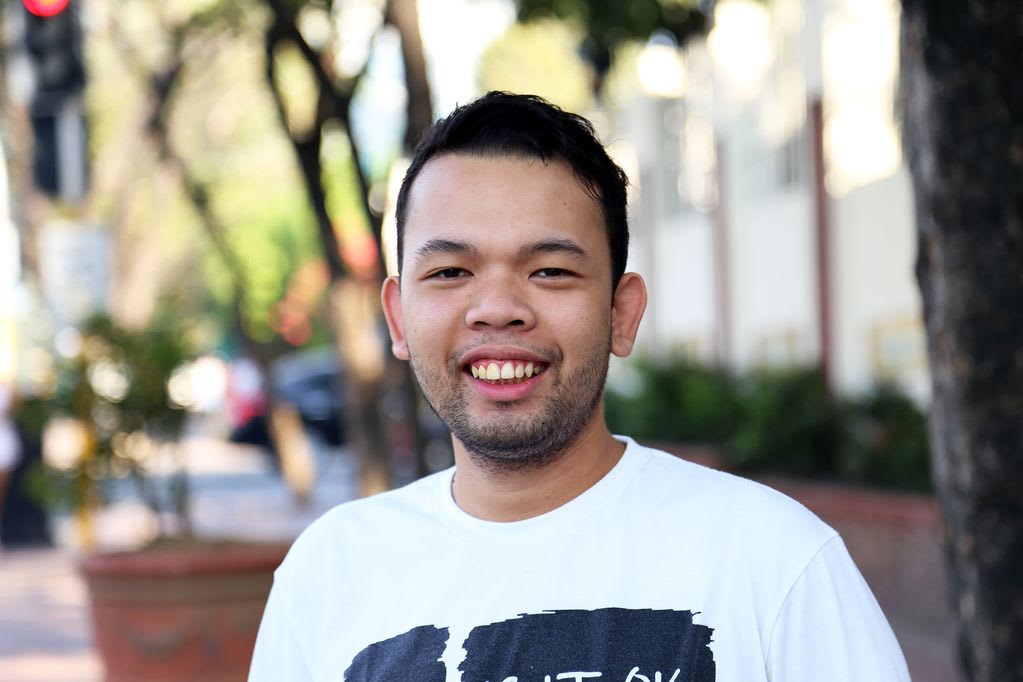 A young man standing outside on a street smiles, wearing a white T-shirt.