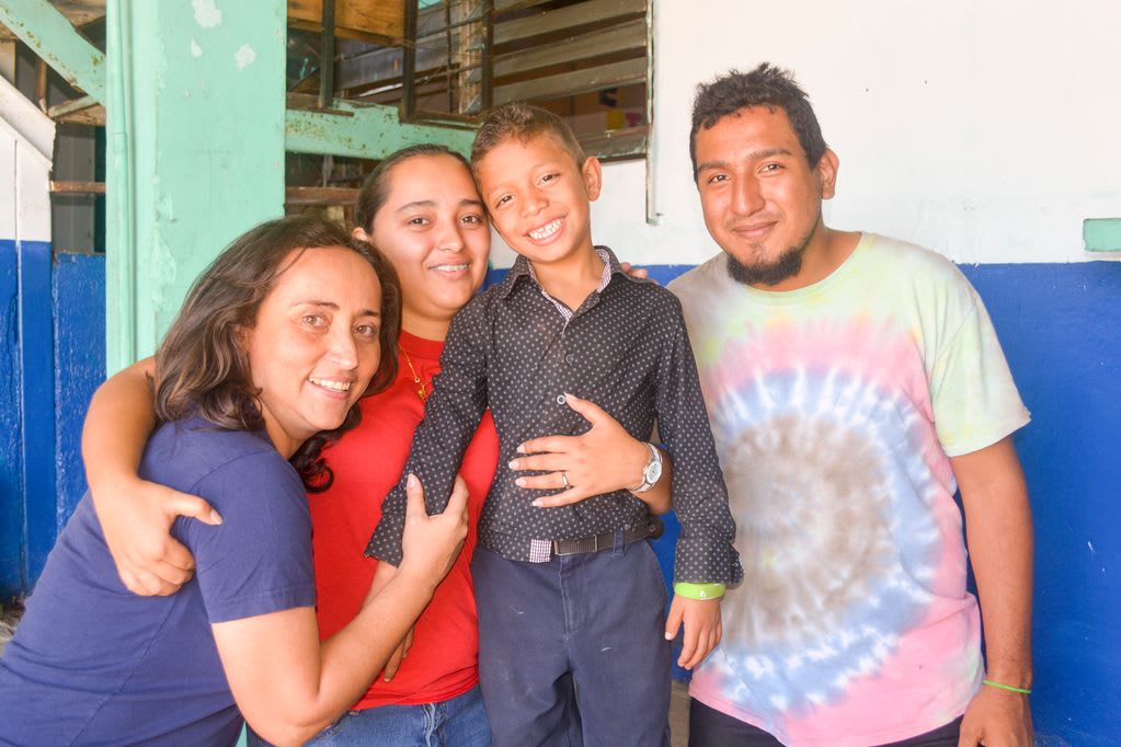 A boy in black is surrounded by three smiling adults, a woman in blue, a woman in red hugging the boy and a man in a tie-dye shirt