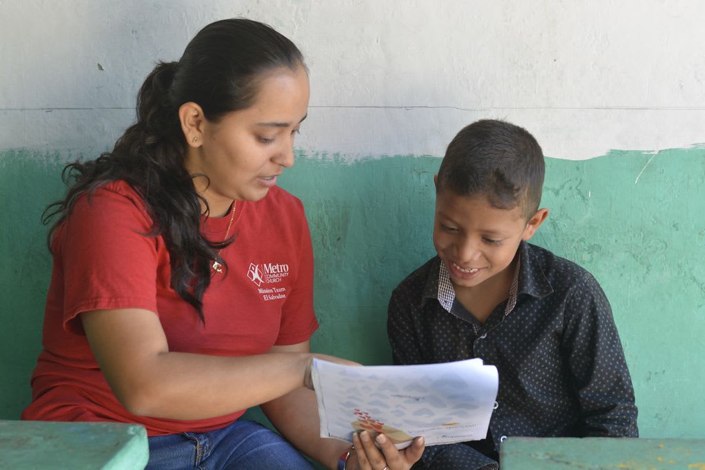 A woman in red sits next to a boy in black pointing at a letter and reading it to him.