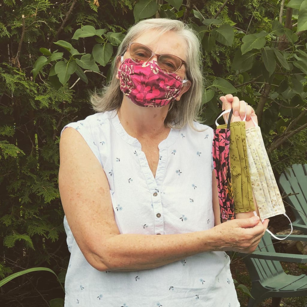 Woman wearing a white shirt wears a pink mask and holds up 3 other masks she has made