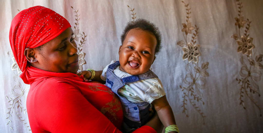 A Ugandan women wearing a red shirt and head dress smiles at the baby she holds in her arms.
