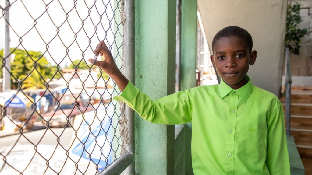 Boy in a green shirt holds onto a fence and looks at the camera