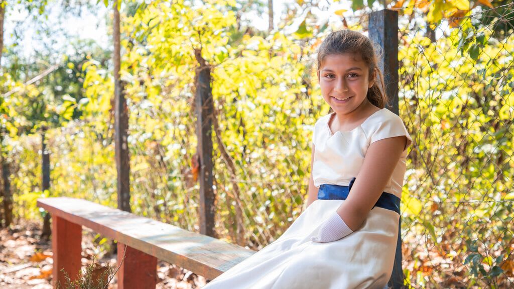 Girl wearing a white dress sits on a bench infront of trees. She wear a sock on her hand. She is beautiful.