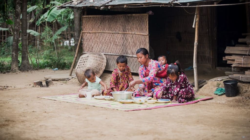 Family sits on a mat outside their home sharing a pot of food.