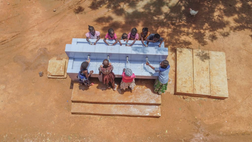A group of children stand a blue sink block and wash their hands.