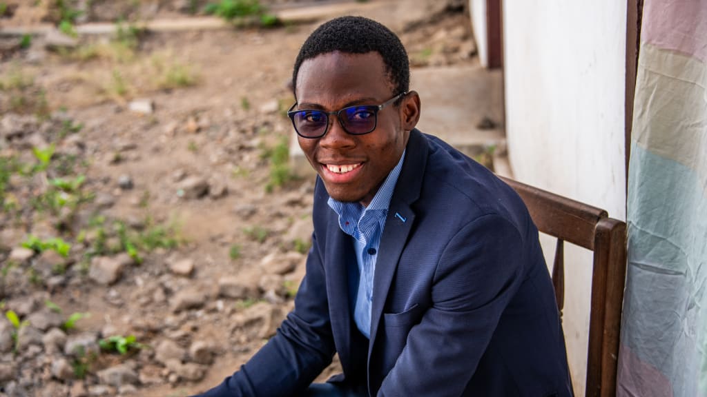 A young man wearing sunglasses and a blue suit sits in front of his childhood home in Togo. He is the CEO of a tech start up where he will work after graduating from the Compassion program.