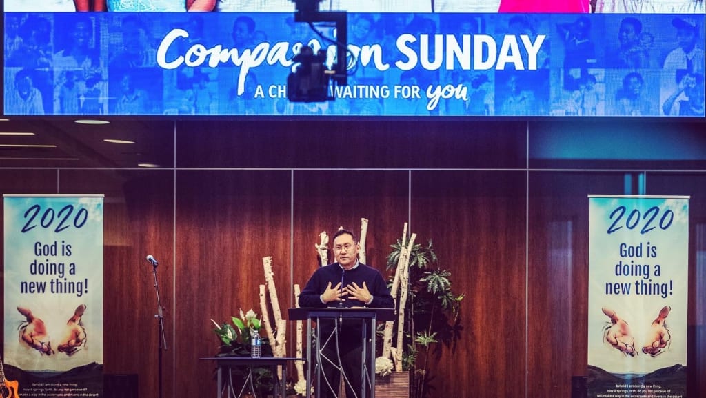A man dressed in black stands behind a pulpit on stage with a blue "Compassion Sunday" banner.