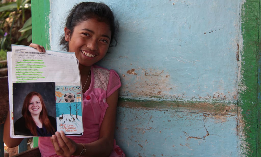 An Indian girl holds a letter up to show the camera. She smiles and leans against a blue wall
