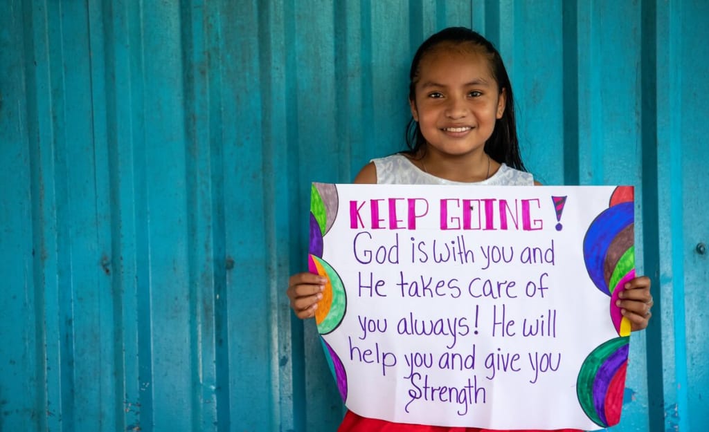 Little girl stands against a turquoise wall holding a sign that says, "Keep going! God is with you and He takes care of you always. He will help you and give you strength."