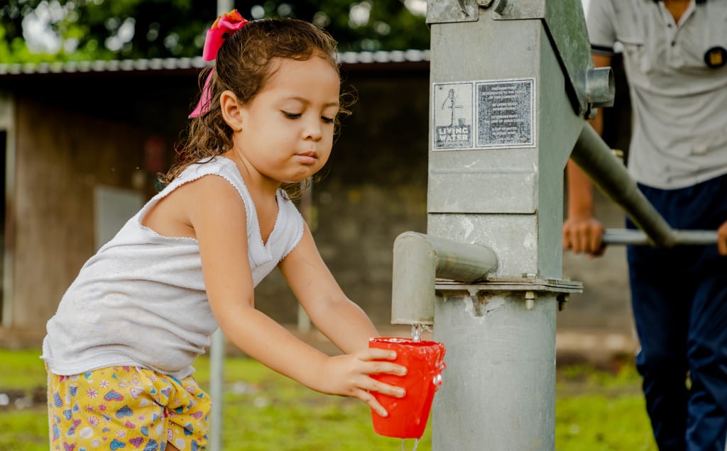 A young girl with a bow in her hair holds a red cup under a water pump.