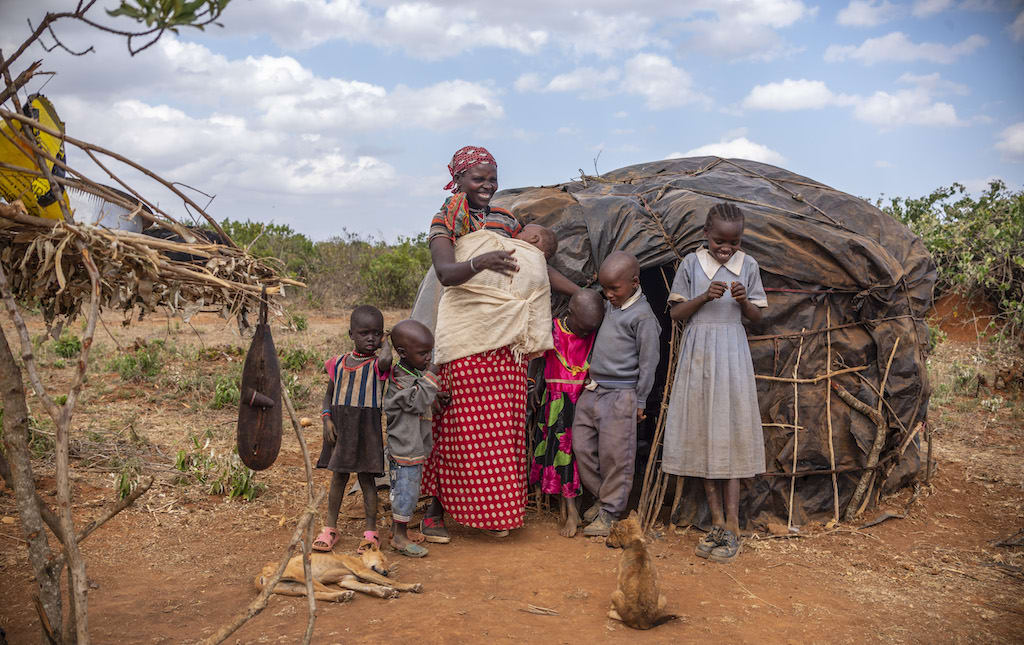 A family of seven people, a mother and six children, stands in front of a small black hut.