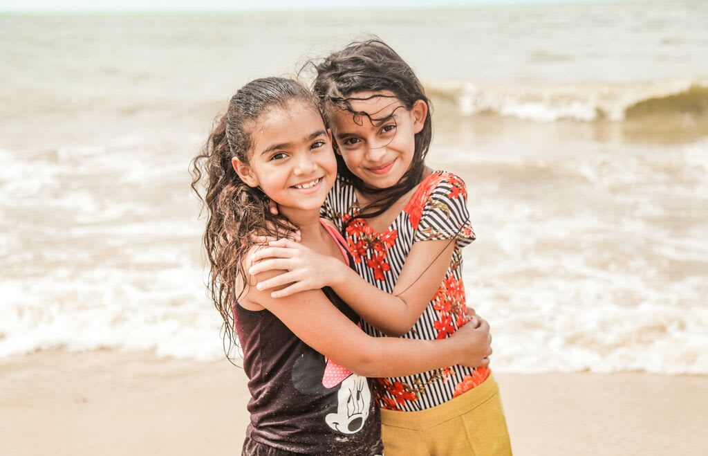 Two little girls hug while looking at the camera, standing in front of the ocean.