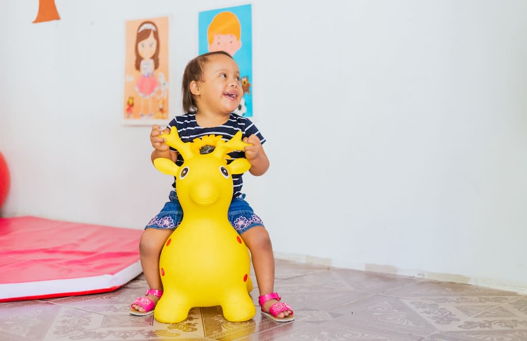 A baby girl sits on a bouncy yellow toy deer. She is laughing and looking to the side.