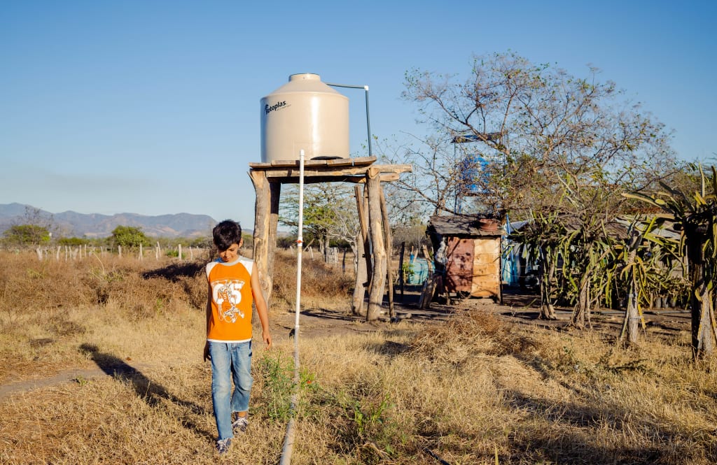 A young boy wearing an orange shirt walks through a dry field with a water tank behind him