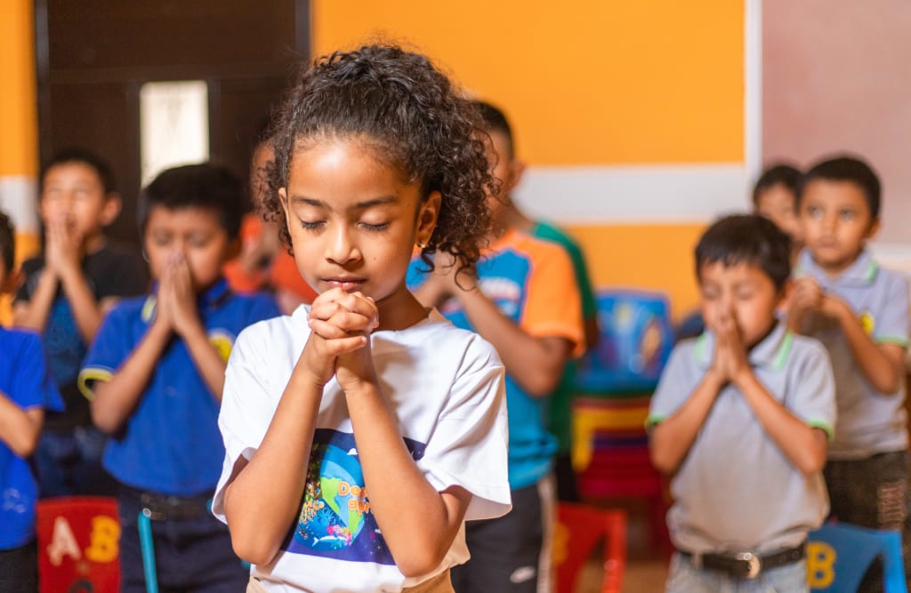 A girl stands with her hands clasped in prayer in a yellow room. There are other children praying behind her.