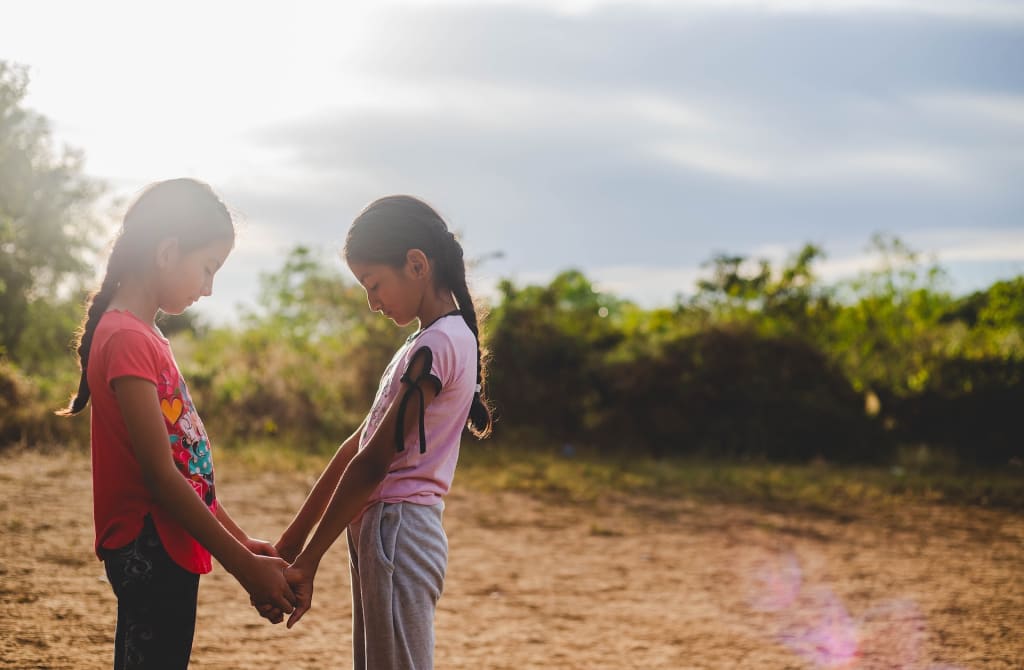 Two young girls hold hands and bow their heads together in prayer.