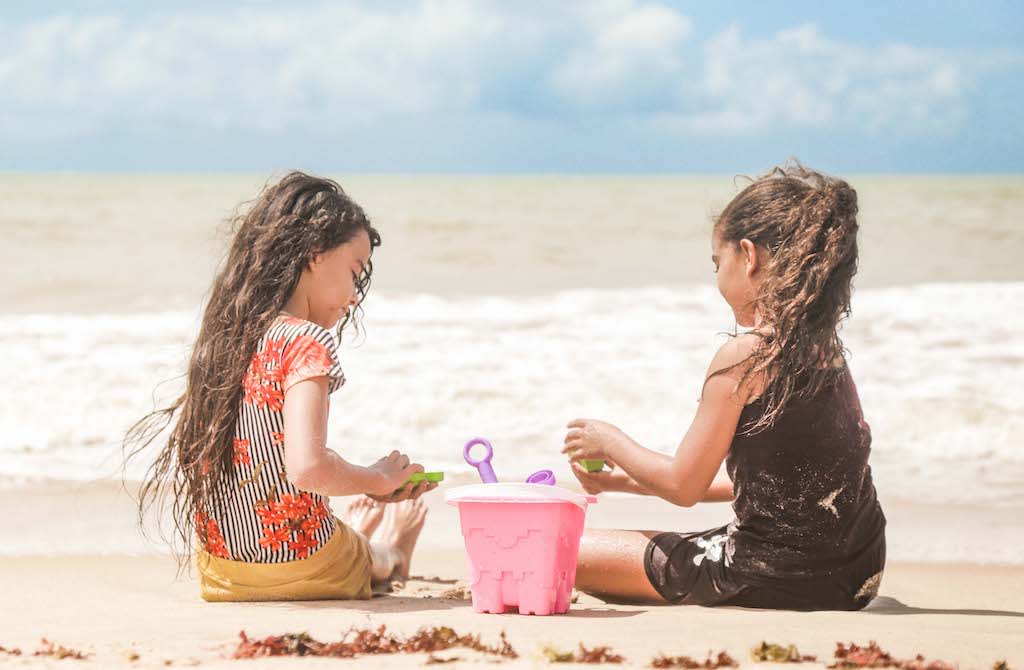 Two young girls play with a pink bucket in the sand at the beach.