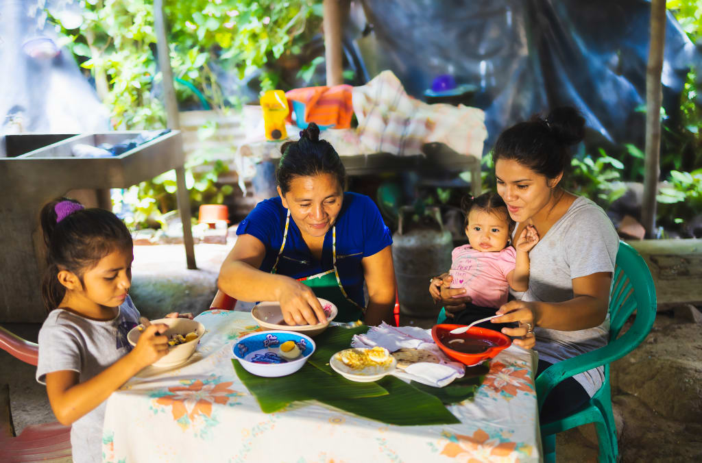 Verenice is wearing a light gray and white dress with a pink and purple pattern on the front. She is sitting at a table with her mother, sister, and niece. They are all eating food that Maritza made using supplies they received from Compassion.
