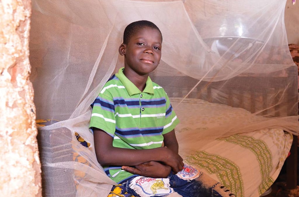 A boy sits on his bed. A mosquito net is draped behind him.