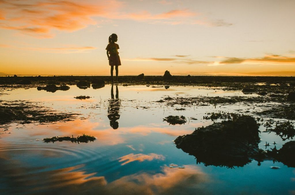 This silhouette photo is of a sunset over the water with a little girl's shadow as she stands by the water.