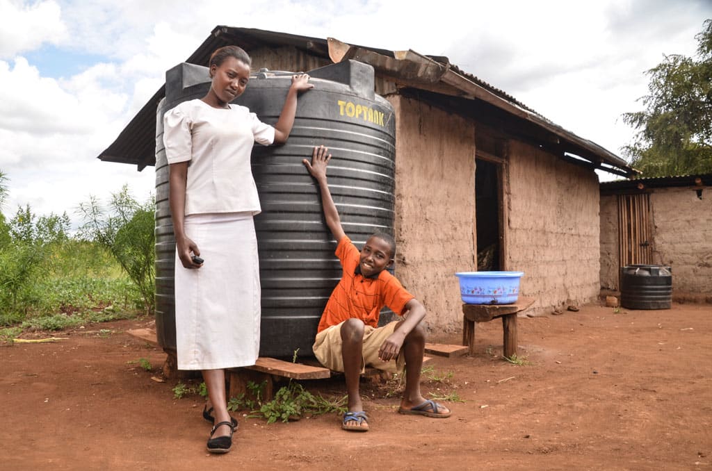 Thirteen-year-old Eric Njuki, a young teen boy, wearing an orange shirt and khaki shorts, at home, with his mother, Rose Nduta, adult female wearing a white shirt and skirt, are standing with a Compassion-provided water tank to harvest rain water, large black water reservoir, container. A walled mud home and greenery is in the background.