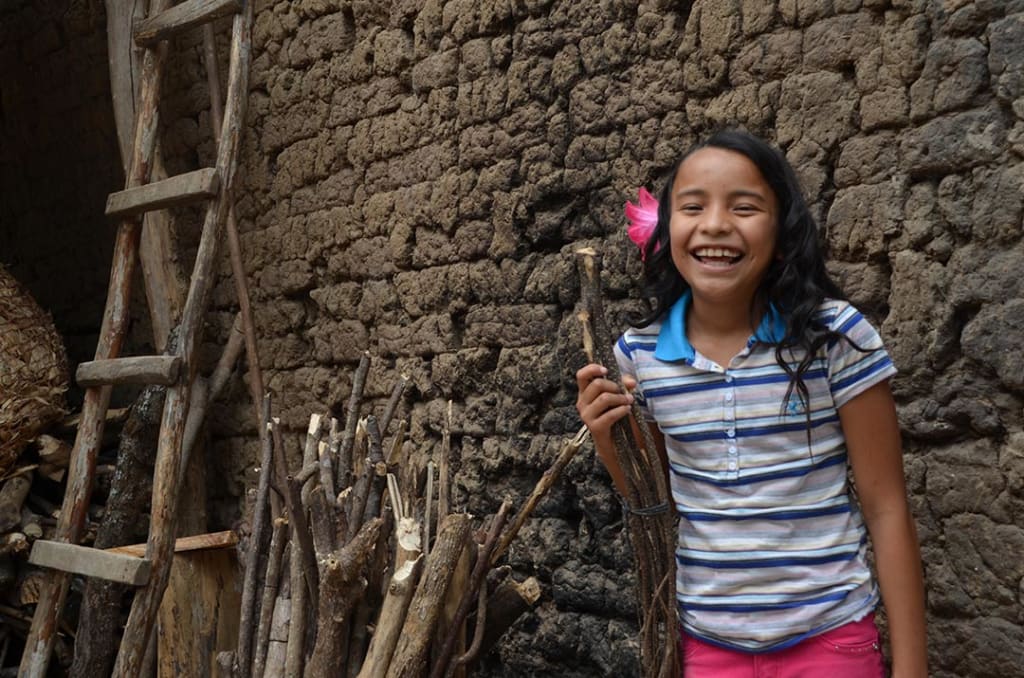 A girl laughs as she stands beside a pile of sticks, leaned against a wall.