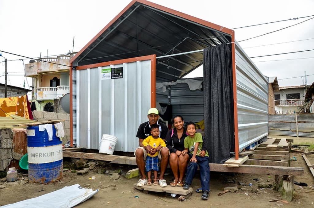 Dastin and his family sit in front of their new home. It's made of corrugated steel and stands on a wooden, raised platform.