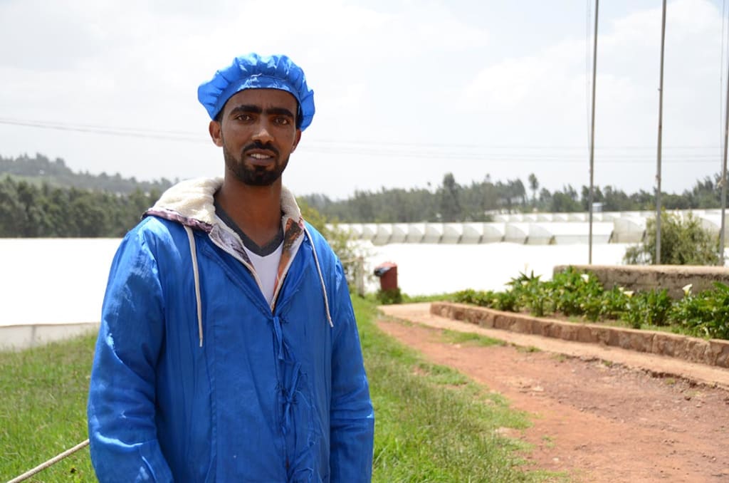 A man stands in front of series of greenhouses. He wears blue hair net and blue, protective lab coat.