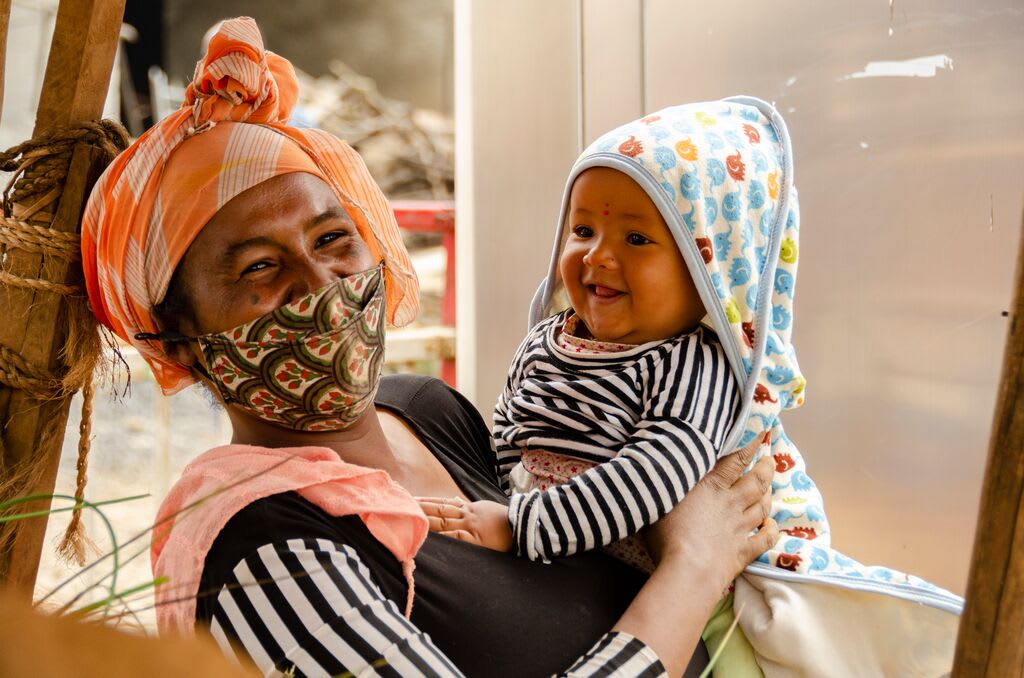 Hanna wears a masks and smiles holding her baby boy, Mikyas, who is wearing a blue hood and a stripped shirt.