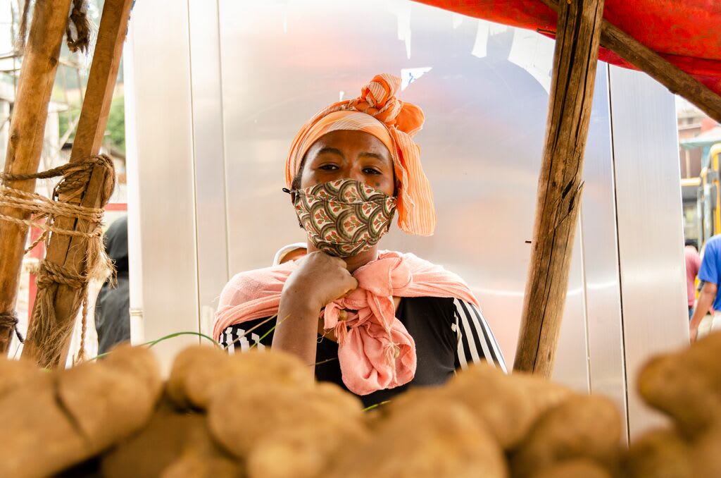 Hanna stands in front of a potato stand, wearing a mask on her face and looking at the camera