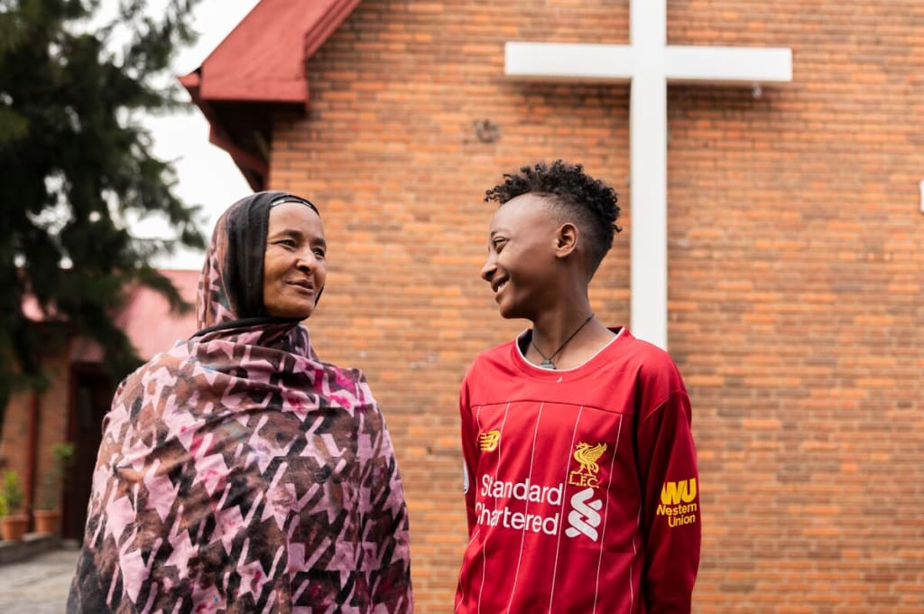 A teen boy is wearing a red shirt. He is standing outside with his mother, who is wearing a pink and brown patterned shawl. They are standing in front of a brick building and talking to each other.