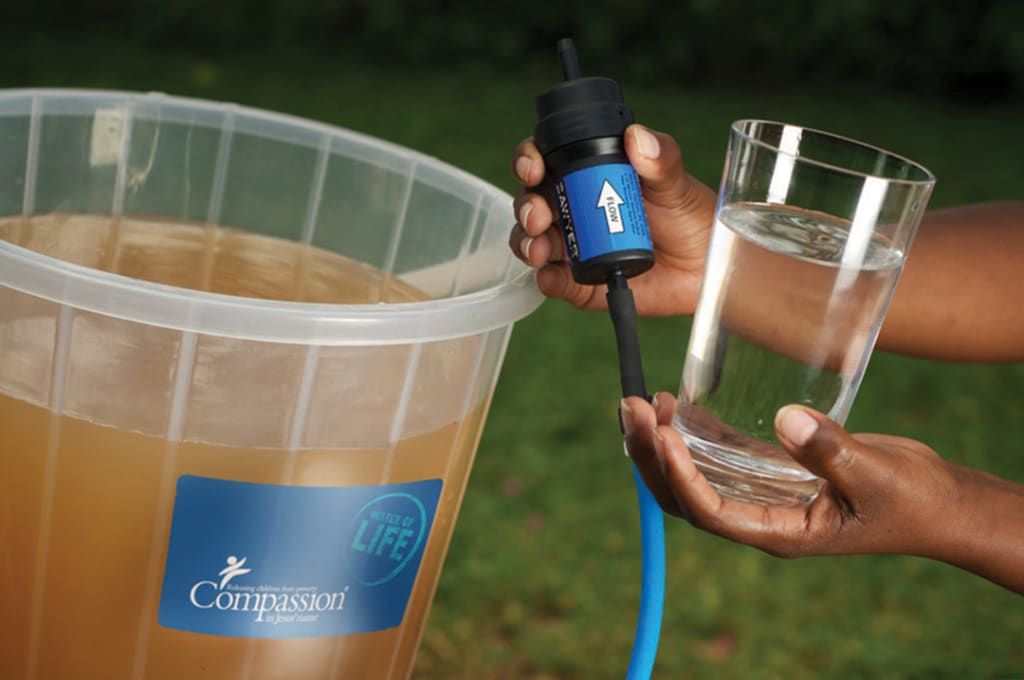 A water filter held up between dirty water and a glass of clean, filtered water.