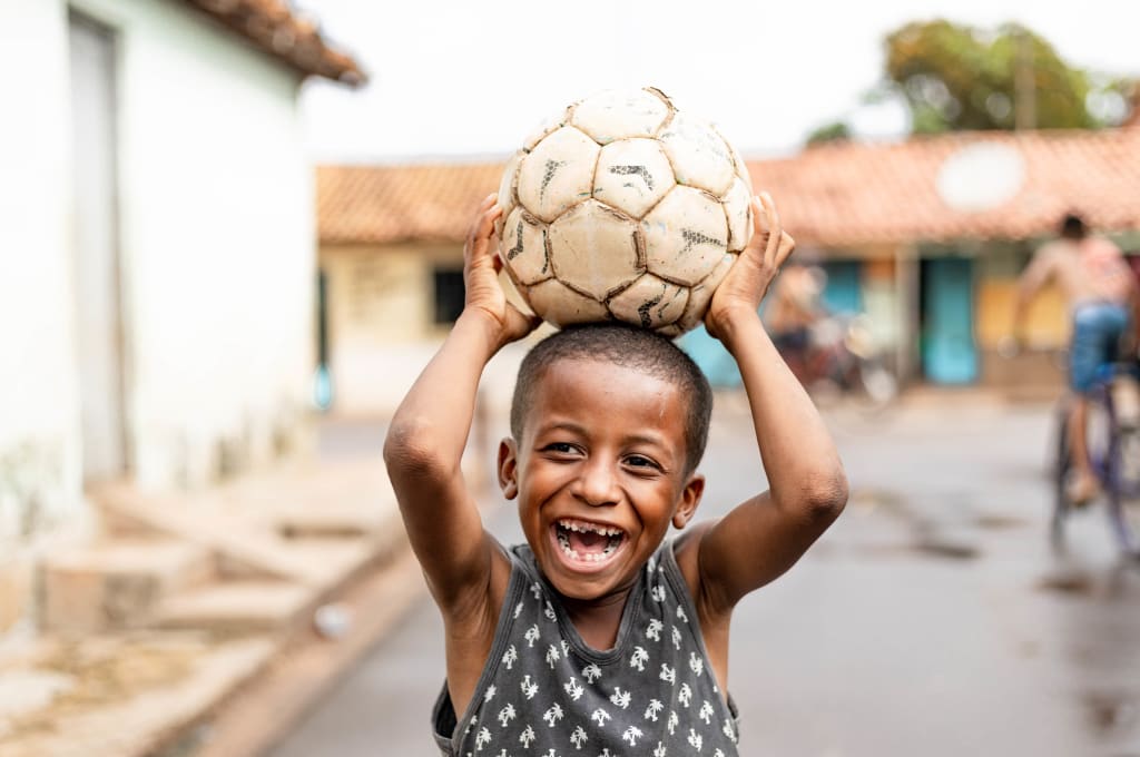 A boy in a grey shirt holds a soccer ball over his head and laughs.
