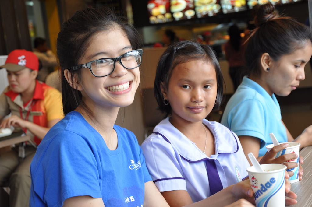A young woman in a blue t-shirt sits next two a young girl in a purple school uniform. They are both looking at the camera.