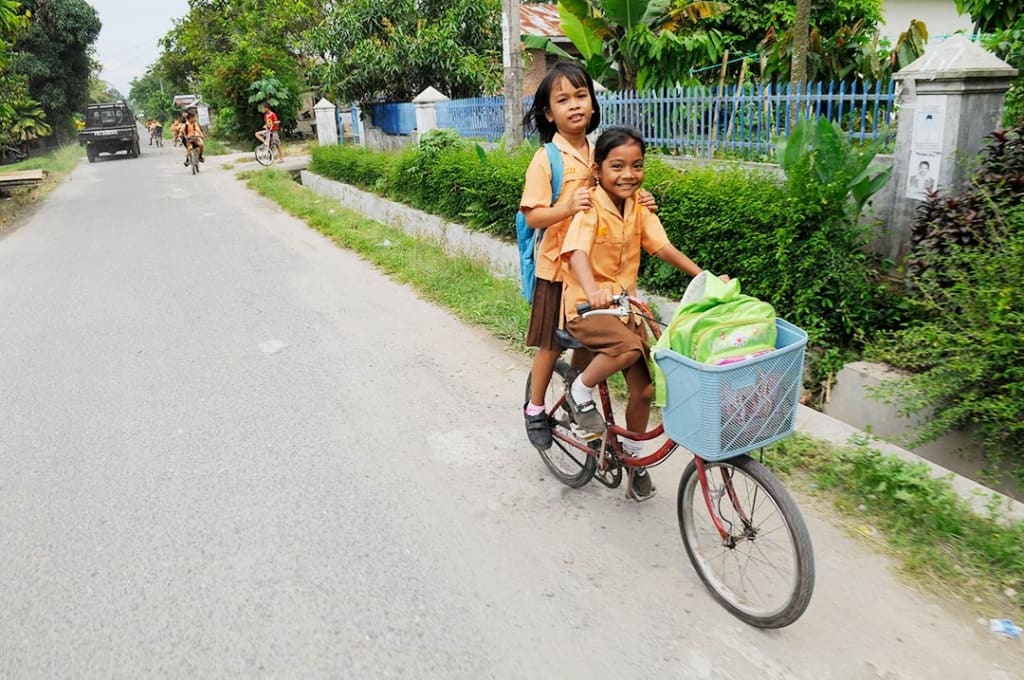 Two girls ride down the street on one bike. They both wear peach coloured shirts with brown dresses.