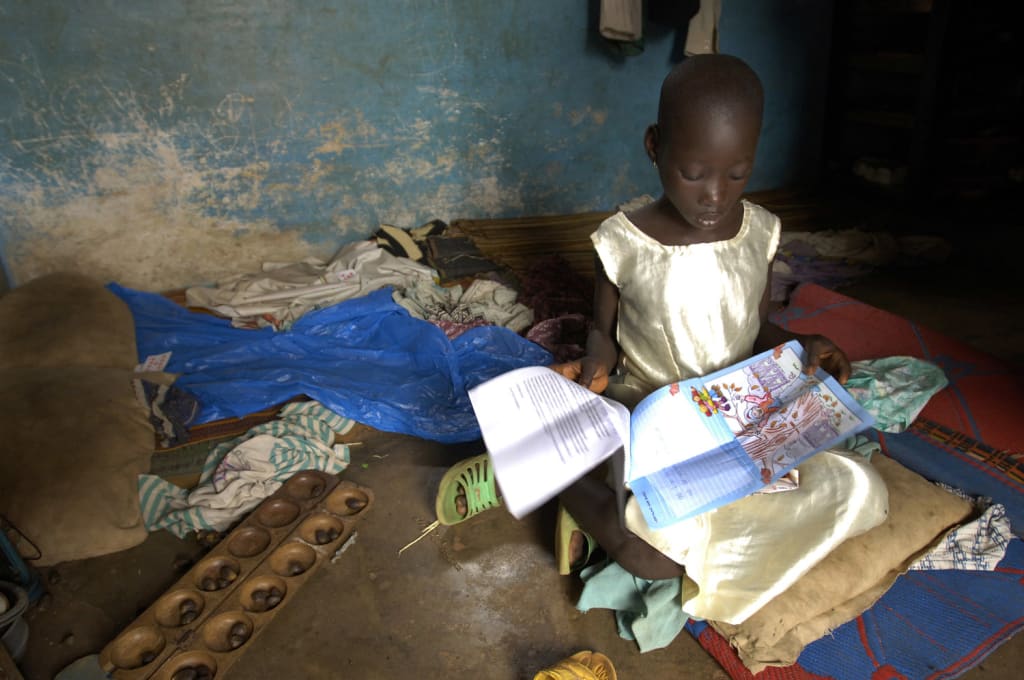 Little girl sits on the ground of her home. She is wearing a yellow dress and reading a letter from her sponsor.