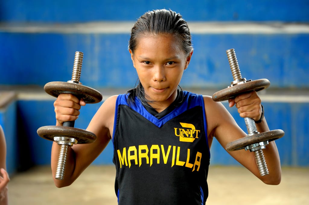 Kenia wears a black and blue jersey and lifts two weights making a tough face