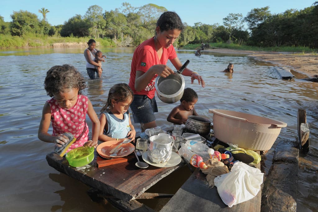 Ana and Thayna Franca de Almeida work with their mother and brother in the water. They are washing plates, pots, and pans in the water. They are using buckets, a bar of soap, and sponge. There is another woman with two other children in the river behind them.