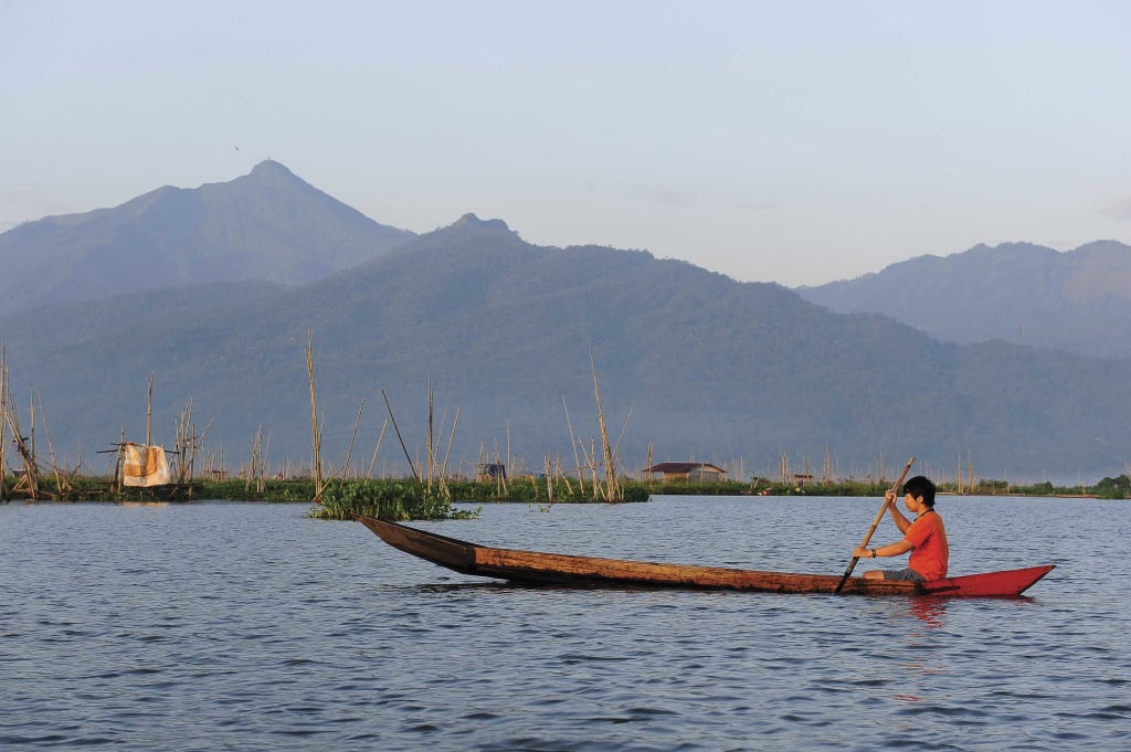 Andistya Han Putra, a teenager, teen, youth, 18 year old project boy, wearing an orange shirt, sits in a canoe, boat, long narrow boat, holding a long bamboo pole in his hands paddling, as the boat floats along river, water. Mountains show in background scenery, landscape.